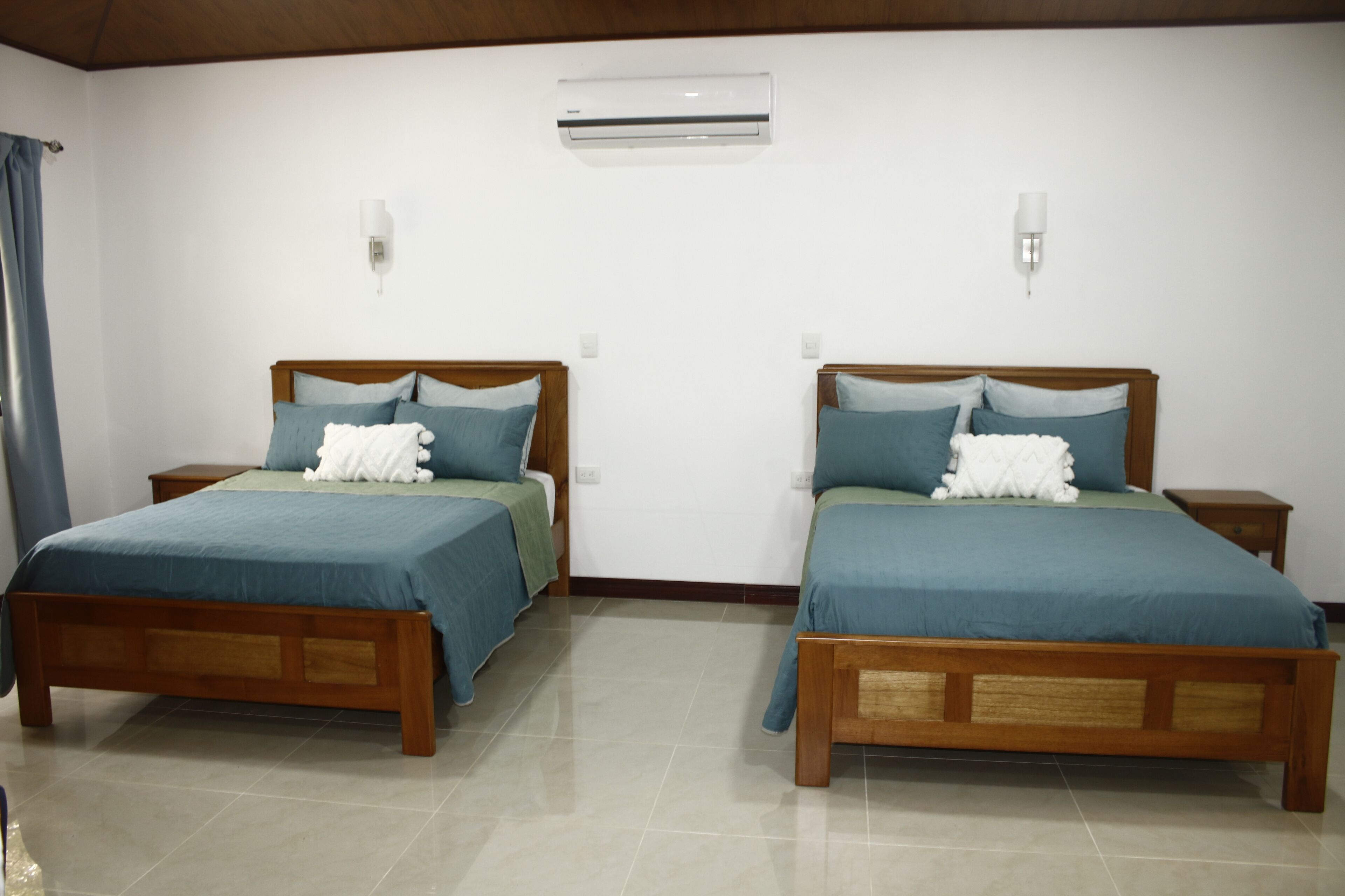 A hotel room with two wooden beds, each with a blue bedspread and pillows, a small bedside table, and an air conditioning unit on the wall.