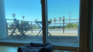 A pet bed and a feeding station by a window overlooking a beach with a wooden deck and palm trees.