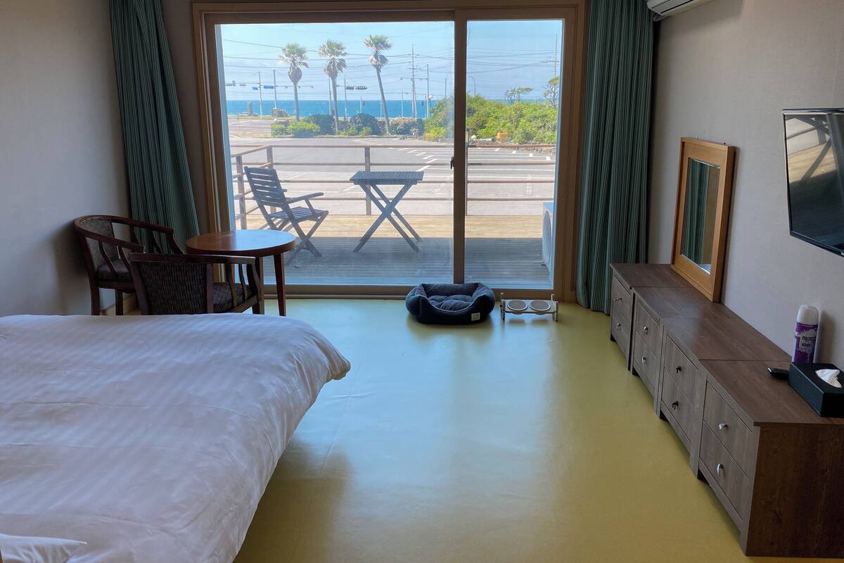 Une chambre d’hôtel comprenant un lit, un bureau, une chaise, une petite table, un téléviseur, une commode et une vue sur la rue et les palmiers, visible à travers une grande fenêtre.