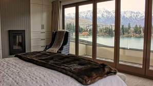Un dormitorio con una cama grande, una chimenea, un sillón con manta y vistas a montañas y un lago a través de puertas correderas amplias.