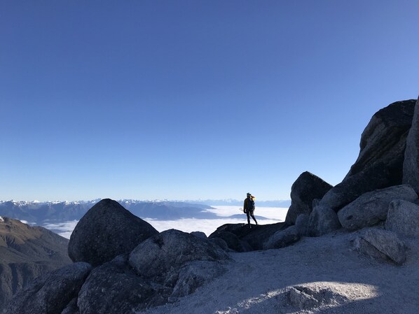 Una persona camina por un sendero rocoso en la montaña, con una vista despejada de montañas nevadas a lo lejos.