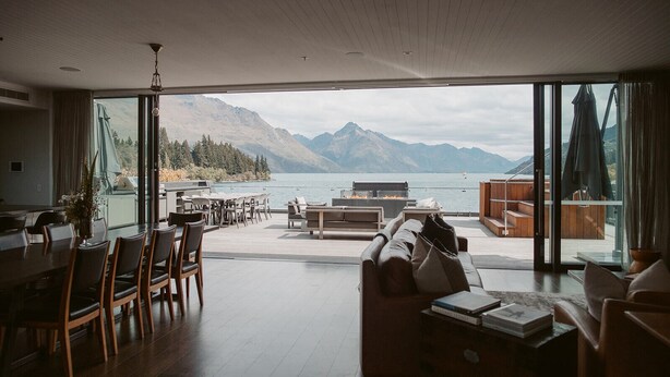 Amplia sala de estar con un ventanal que da a un lago y montañas. Hay un comedor con sillas y una mesa, y una zona de estar con sofás y una mesita de centro.