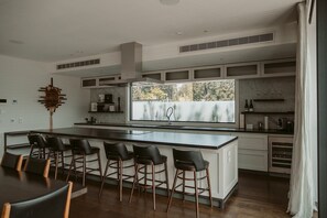 A modern kitchen with a large island, bar stools, and a view of trees through the window.