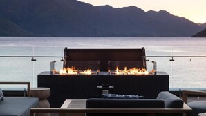 Un área de estar exterior moderna con una chimenea integrada, con vistas a un cuerpo de agua tranquilo y montañas.