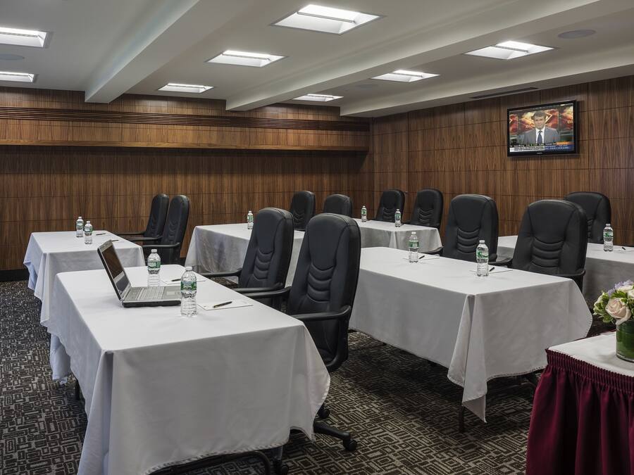 Een vergaderruimte met meerdere tafels, elk voorzien van een laptop, een waterfles en een glas. De ruimte heeft een wand van houten panelen en een televisie die daaraan bevestigd is.