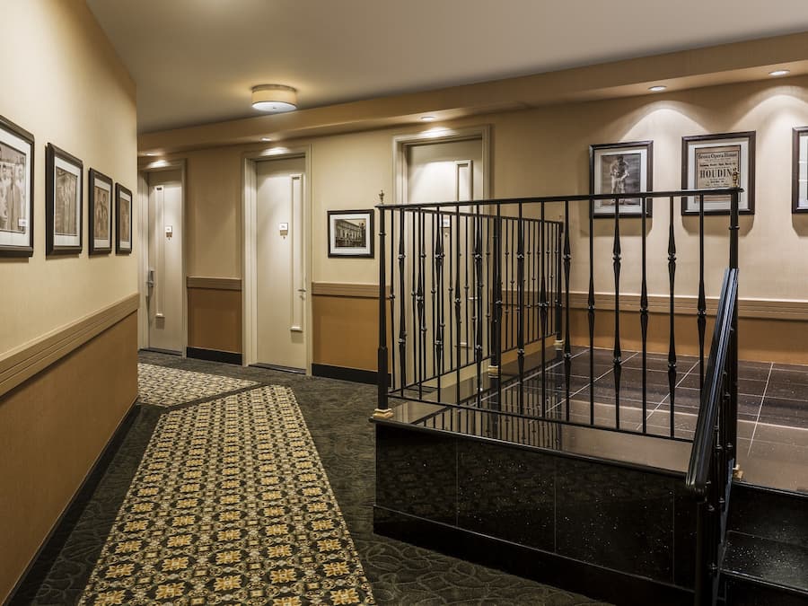 A hotel hallway with framed pictures on the walls, a patterned carpet, and a staircase with a metal railing.