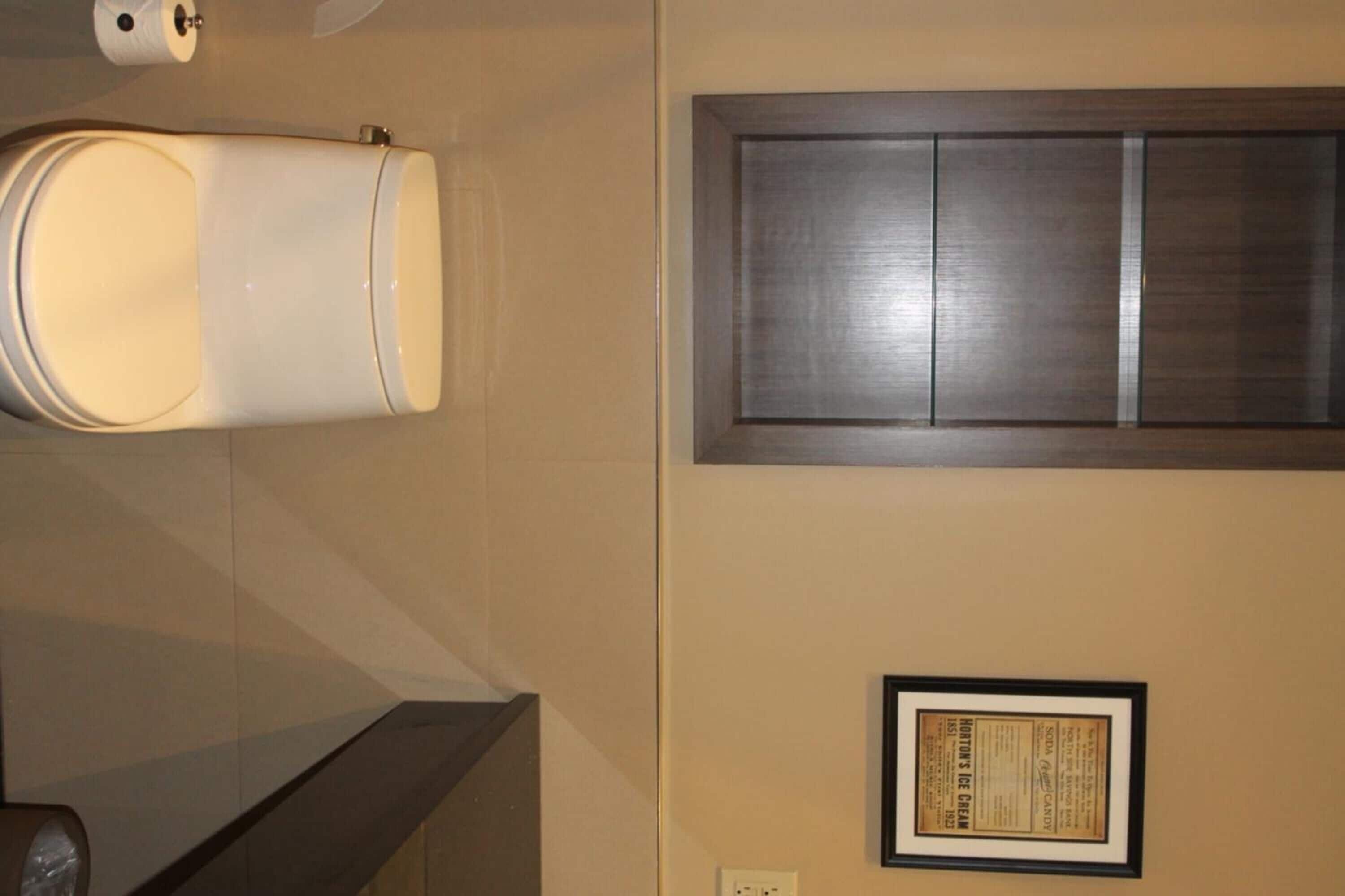 A restroom with a toilet, a trash can, a framed sign on the wall, and a cabinet above the toilet.