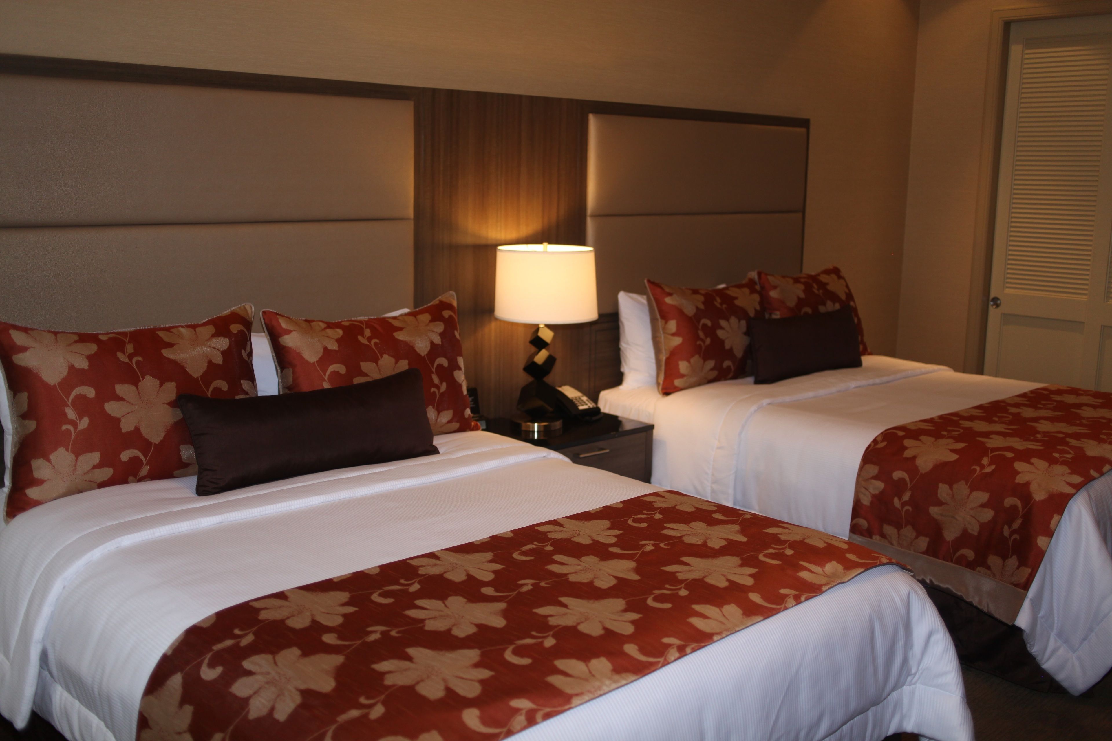 A hotel room with two beds, each with a red floral bedspread and matching pillows. There is a nightstand with a lamp and a telephone between the beds.