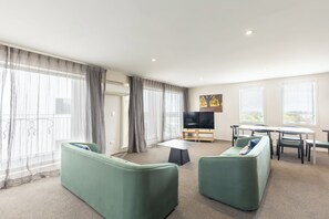 Ein modernes Wohnzimmer mit grünen Sofas, einem Flachbildfernseher, einem Esstisch und großen Fenstern mit Vorhängen.