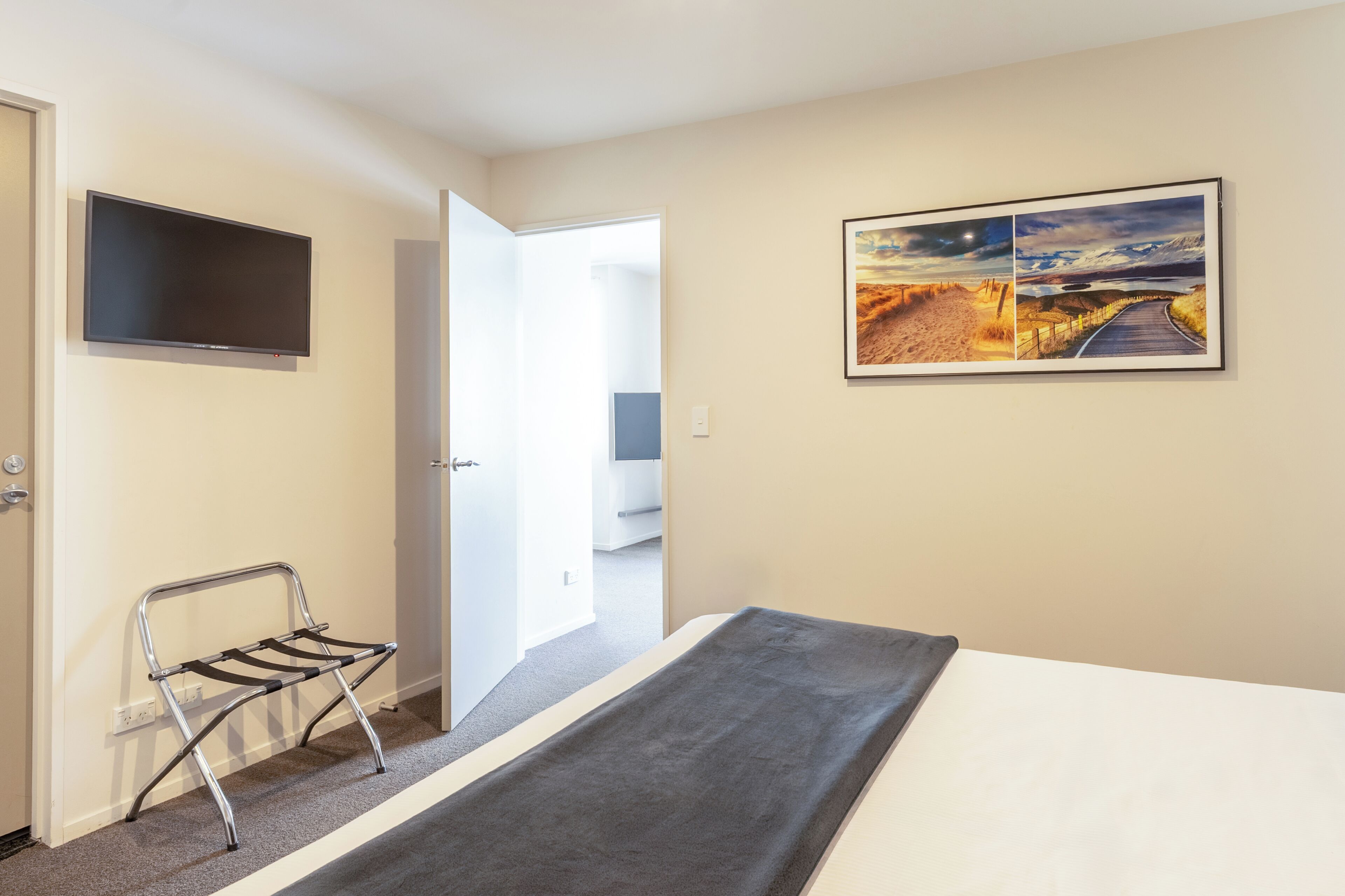 Habitación de hotel con cama, televisor de pantalla plana, banca metálica, cuadro de paisaje enmarco y puerta abierta que da a otra habitación.