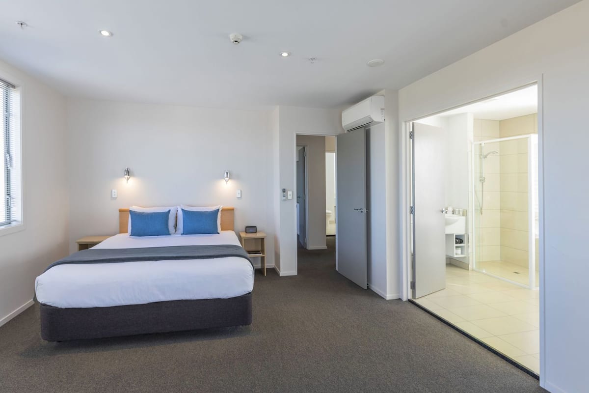 Una habitación de hotel moderna con una cama grande, mesitas de noche, un escritorio, un armario y una zona de ducha con mampara de cristal.