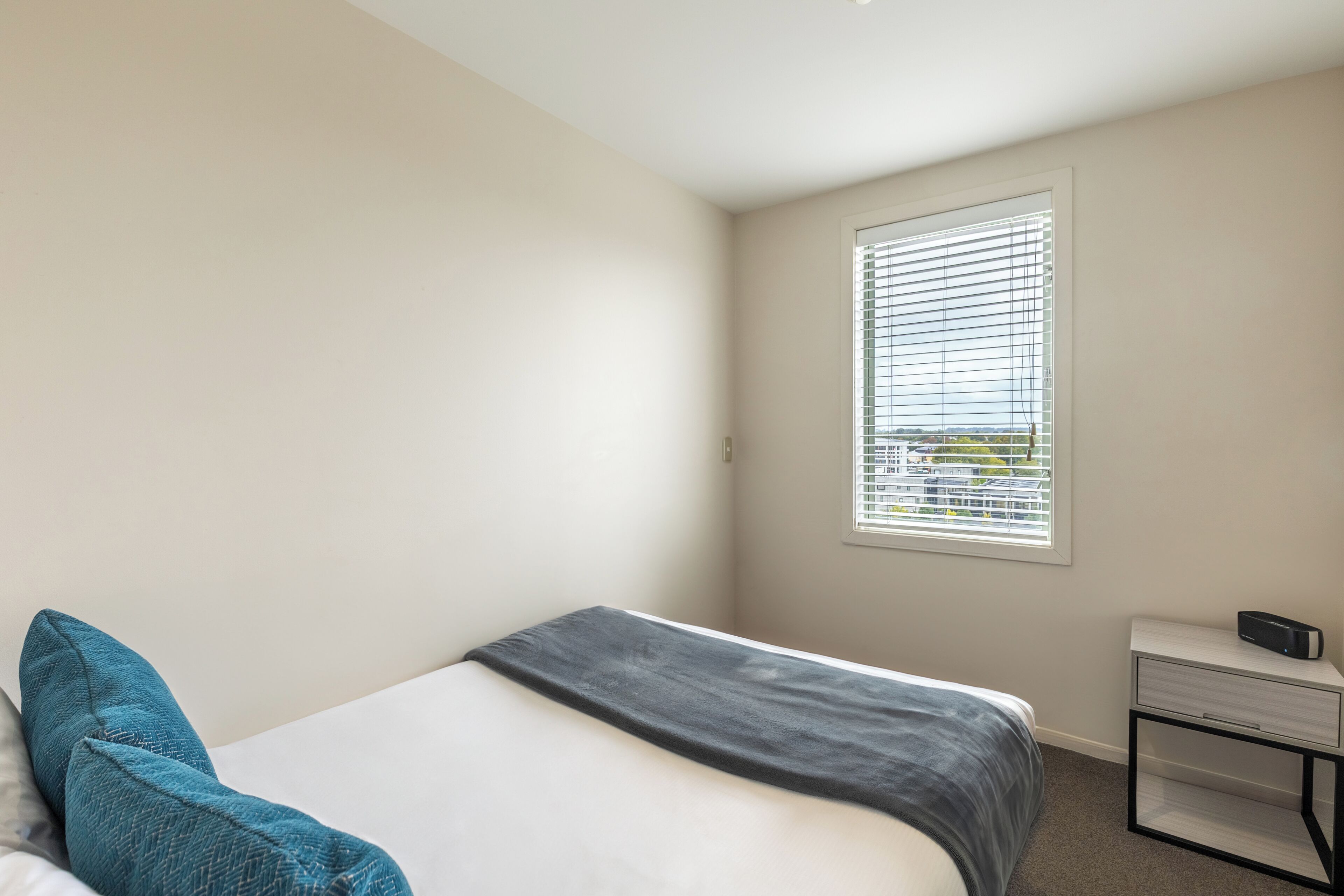 Een slaapkamer met een bed, een nachtkastje, een raam met jaloezieën en uitzicht op buiten.