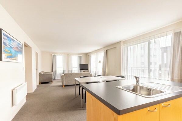 A modern hotel room with a kitchenette, a sofa, a TV, and large windows.