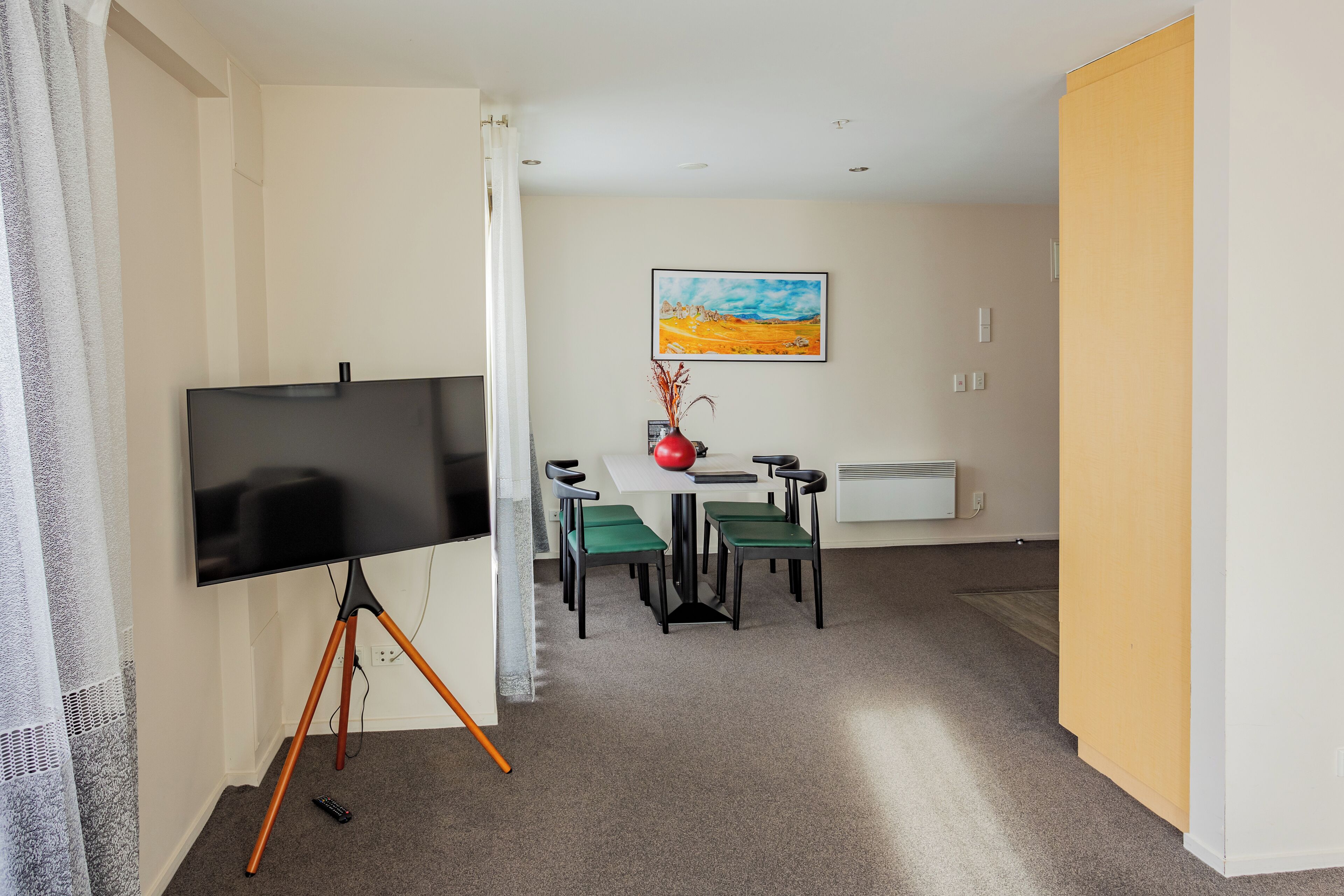 A room with a flat-screen TV on a tripod stand, a dining table with four chairs, a vase with dried plants, and a painting on the wall.