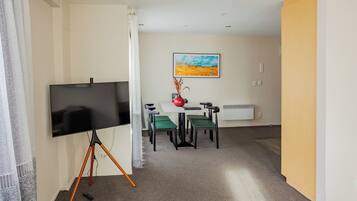 A room with a flat-screen TV on a tripod stand, a dining table with four chairs, a vase with dried plants, and a painting on the wall.