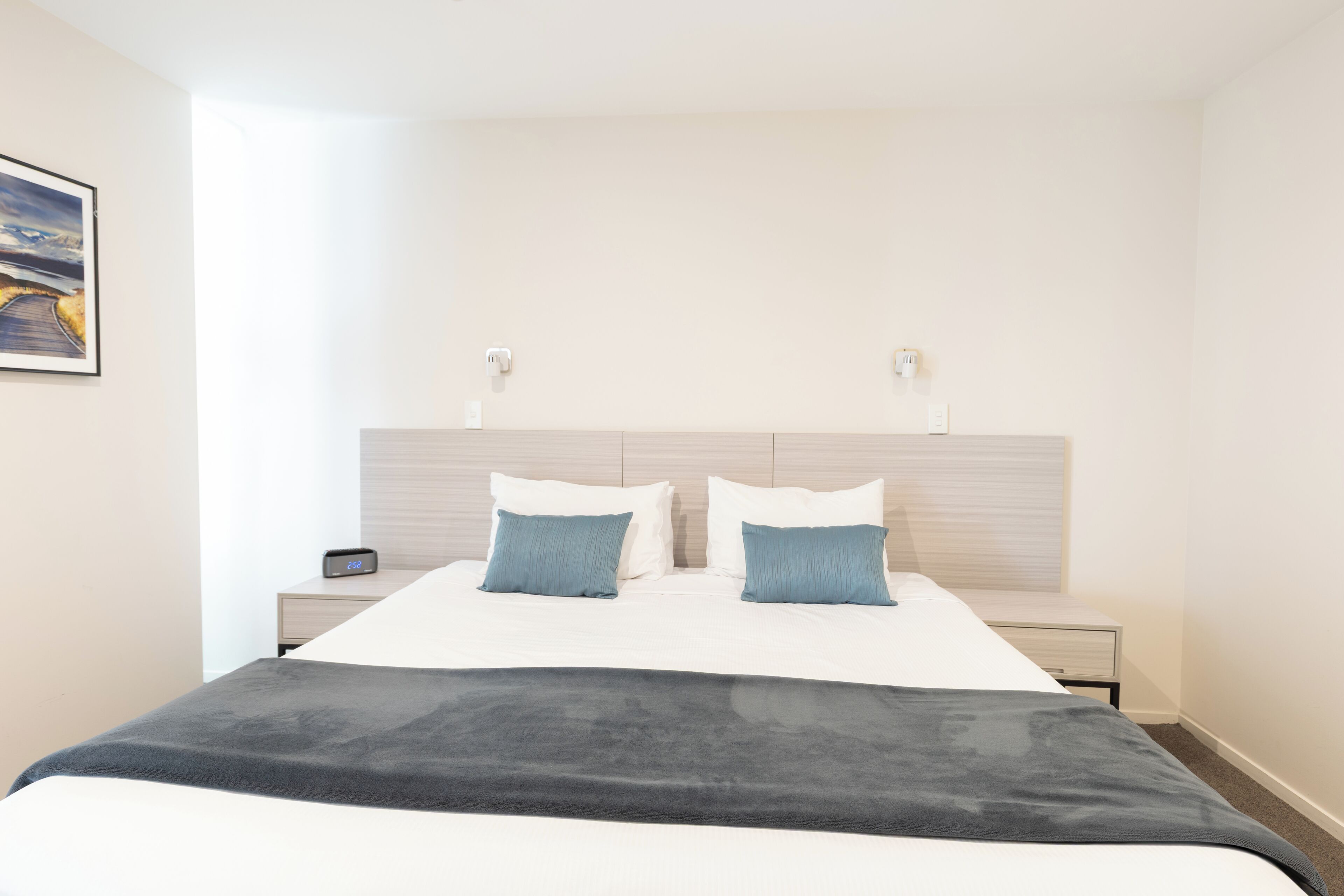 Habitación de hotel con una cama, dos mesitas de noche, una lámpara de pared y un cuadro enmarcado.