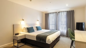 Een hotelkamer met een bed, nachtkastje, lamp, gordijnen en een bureau met stoel.