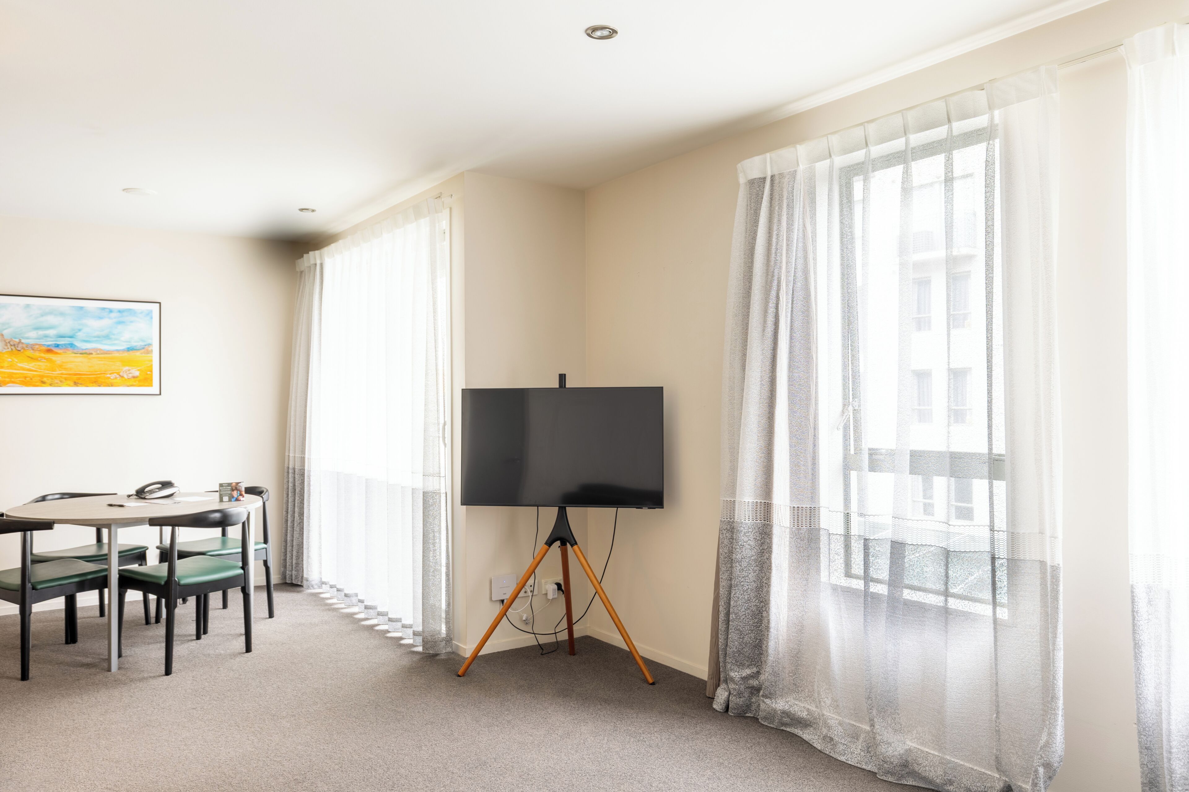 A room with a large flat-screen TV on a tripod stand, a dining table with chairs, and sheer curtains on the windows.