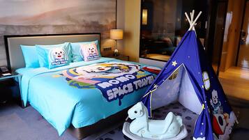 Habitación de hotel con una cama que tiene una colcha con temática de 'Momo Space Travel', un pequeño juguete con forma de caballo blanco en el suelo y una estructura azul tipo tienda con un personaje de dibujos animados.