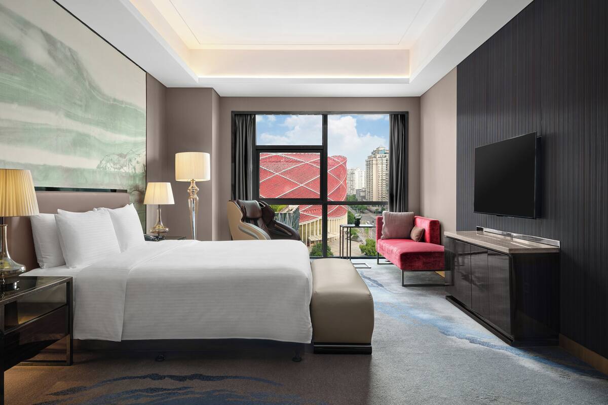 Ein Hotelzimmer mit Bett, Fernseher, einem roten Sessel und Blick auf eine Stadtlandschaft durch große Fenster.