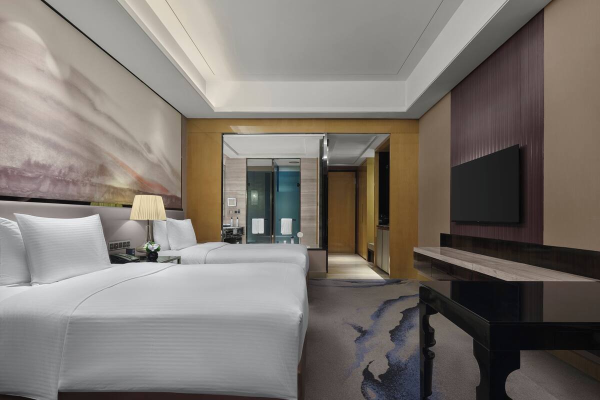 Ein Hotelzimmer mit einem großen Bett, einem Schreibtisch, einem Flachbildfernseher und einem durch eine Glastür einsehbaren Badezimmer.