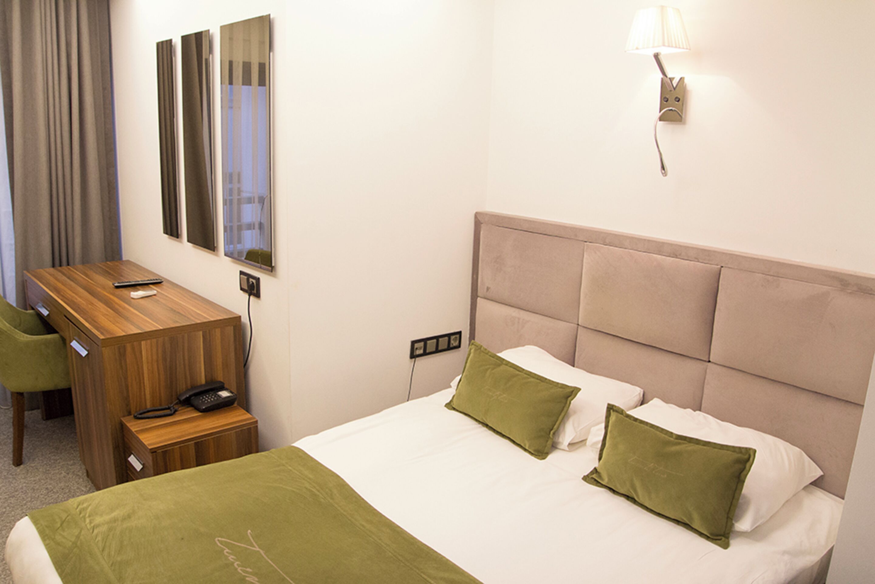 Ein Hotelzimmer mit Bett, grünen Kissen, einem Holztisch mit Telefon und einem Spiegel an der Wand.