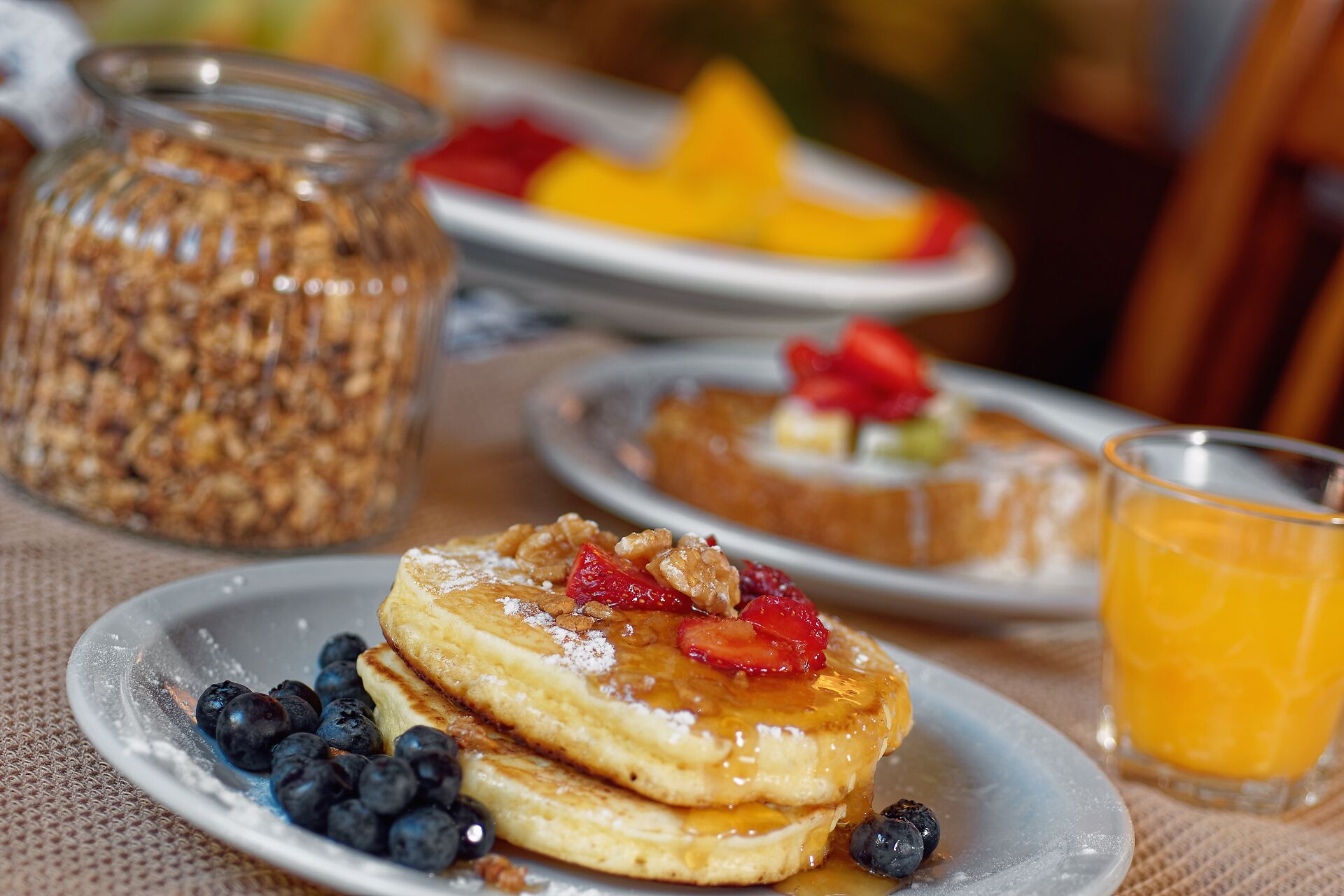 Une table dressée pour le petit-déjeuner avec des crêpes garnies de fraises et de myrtilles, un verre de jus d’orange et un bocal de granola.