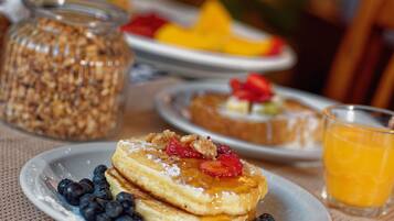 Une table dressée pour le petit-déjeuner avec des crêpes garnies de fraises et de myrtilles, un verre de jus d’orange et un bocal de granola.