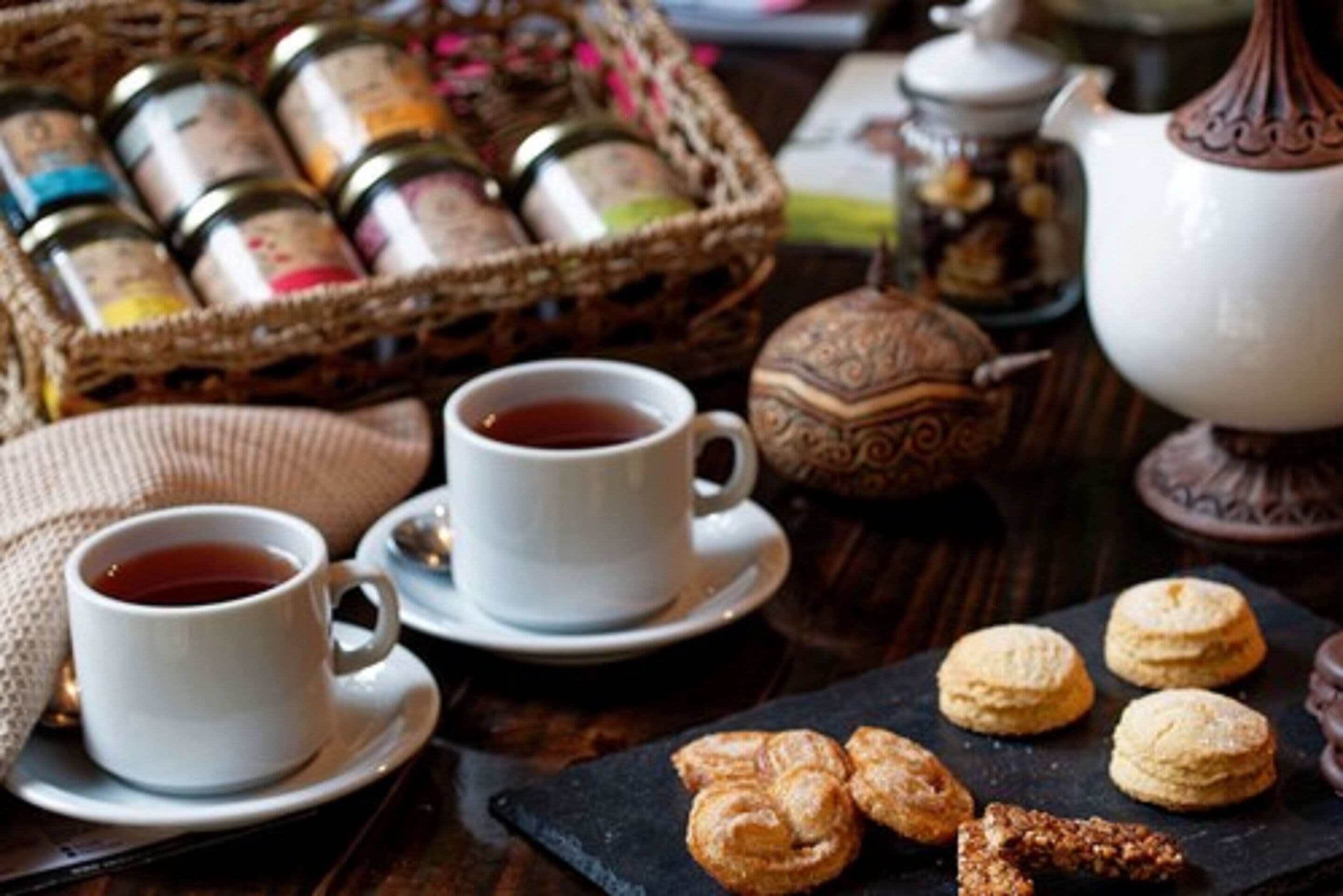 Una mesa con dos tazas blancas de té, una cesta de frascos, un hervidor y un plato de pasteles.