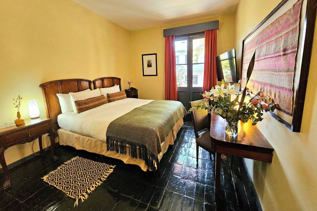Habitació d'hotel amb un llit, una tauleta de nit, un escriptori, una cadira, una finestra amb cortines i un quadre a la paret.