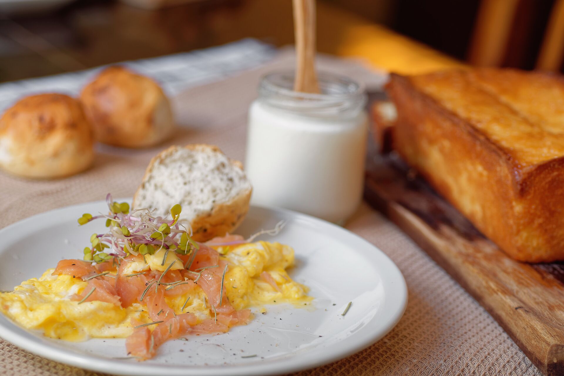 A breakfast plate with scrambled eggs, smoked salmon, and a small salad, accompanied by a glass of milk, a slice of bread, and two rolls.