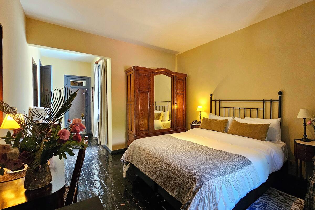 Habitació d'hotel amb llit, taula de nit, làmpada, cadira i mirall.