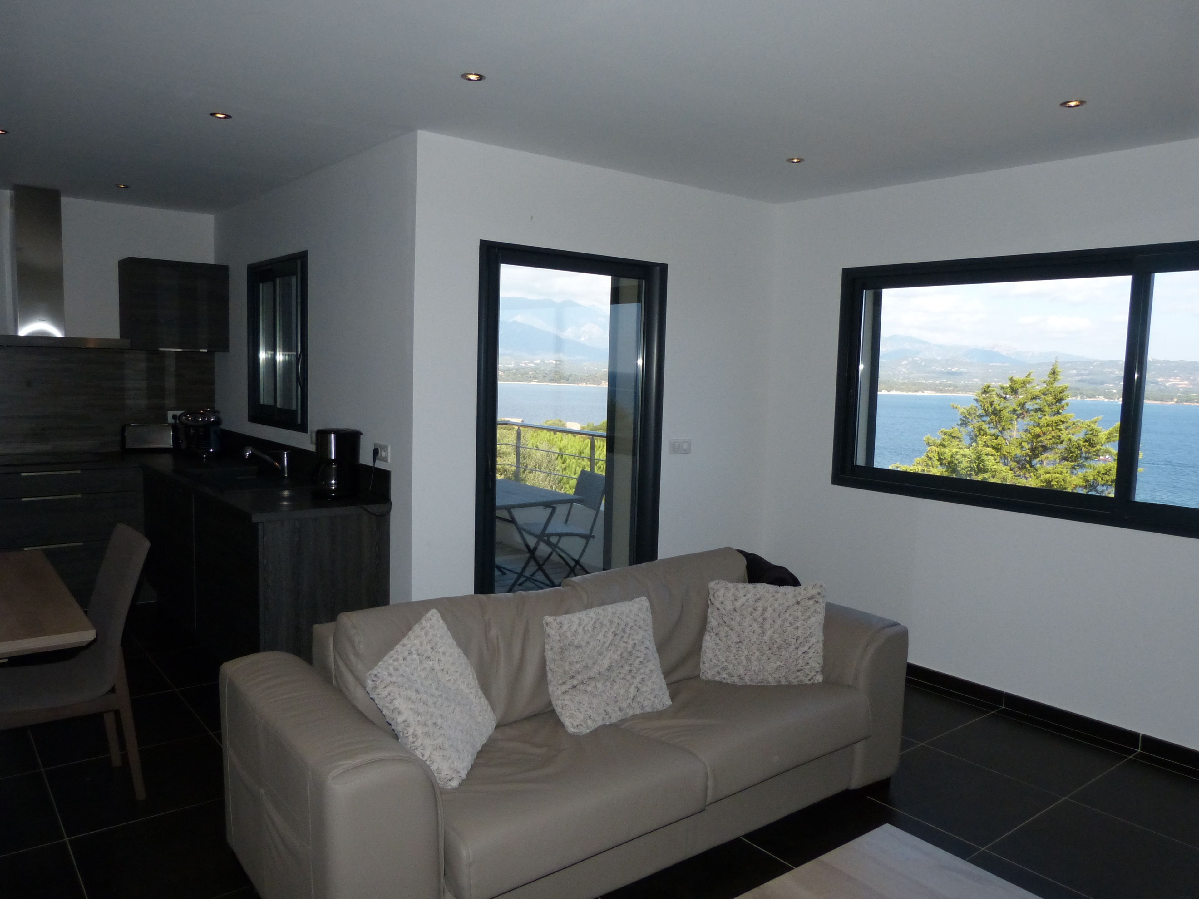 A modern living room with a sofa, a dining area, and a kitchen. There is a large window offering a view of the sea and mountains.