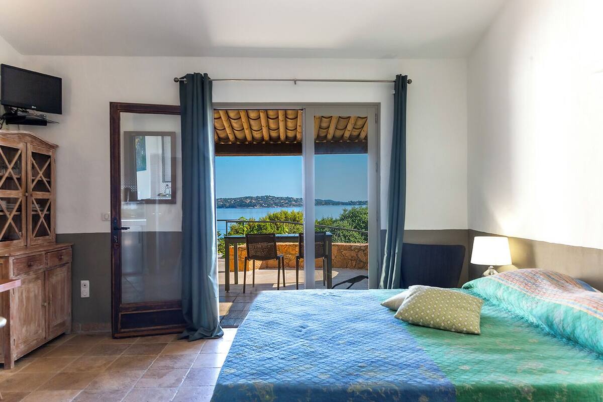 A hotel room with a bed, a TV, a wooden cabinet, and a balcony with a view of the sea.