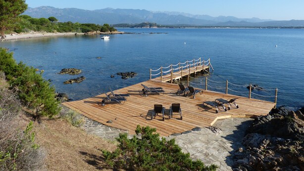 Une jetée en bois, agrémentée de chaises et d’une rambarde, surplombe un lac paisible, entouré de montagnes et de verdure.