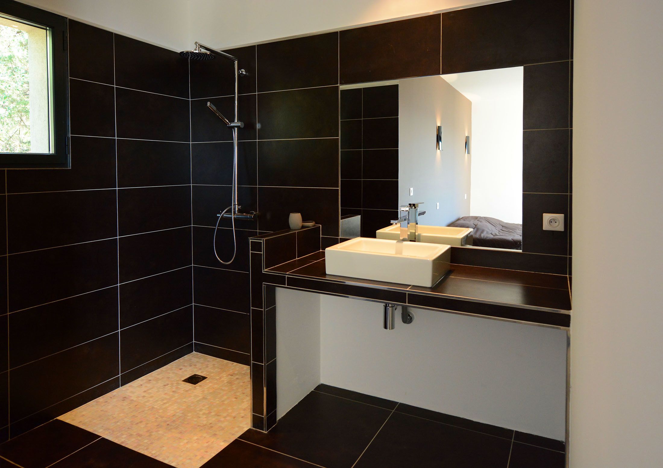A modern bathroom with black tiles, a walk-in shower, a large mirror, and a beige rug.