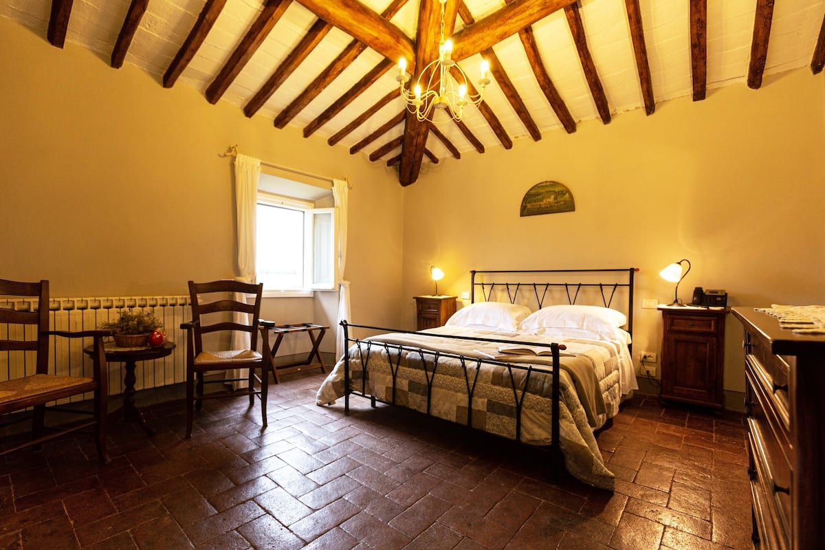 Een slaapkamer met een houten plafond, een bed met metalen frame, een stoel, een tafel met een plant en een raam.