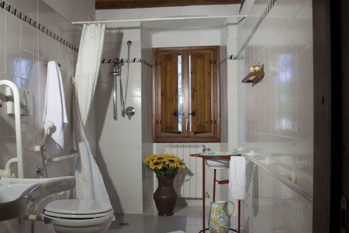 Un baño con techo de madera, baldosas blancas, ducha, inodoro, lavamanos y ventana.