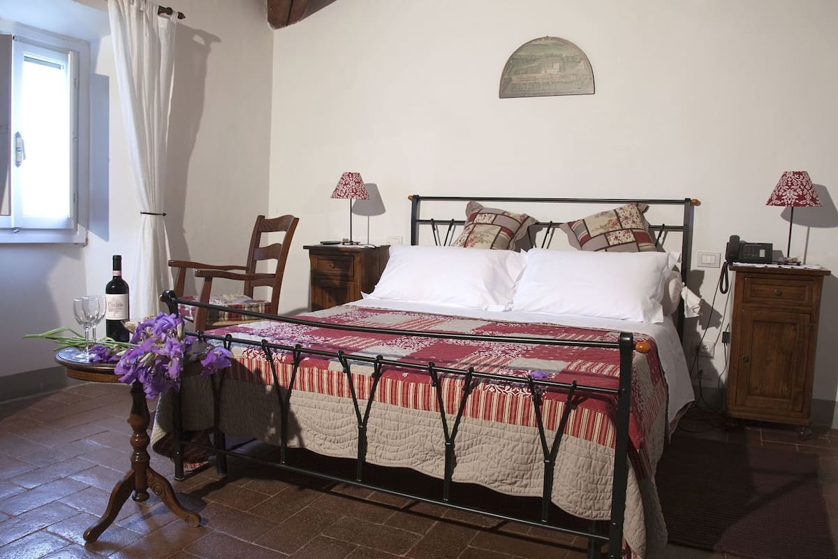 Een slaapkamer met een bed, een stoel, een nachtkastje, een lamp, een fles wijn en een vaas met bloemen.