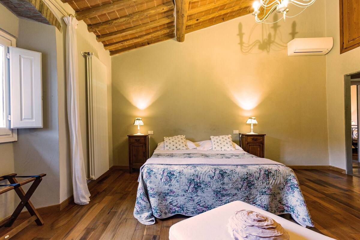 Een slaapkamer met een houten plafond, een bed met een bloemenbedekking, twee nachtkastjes met lampen en een raam met gordijnen.