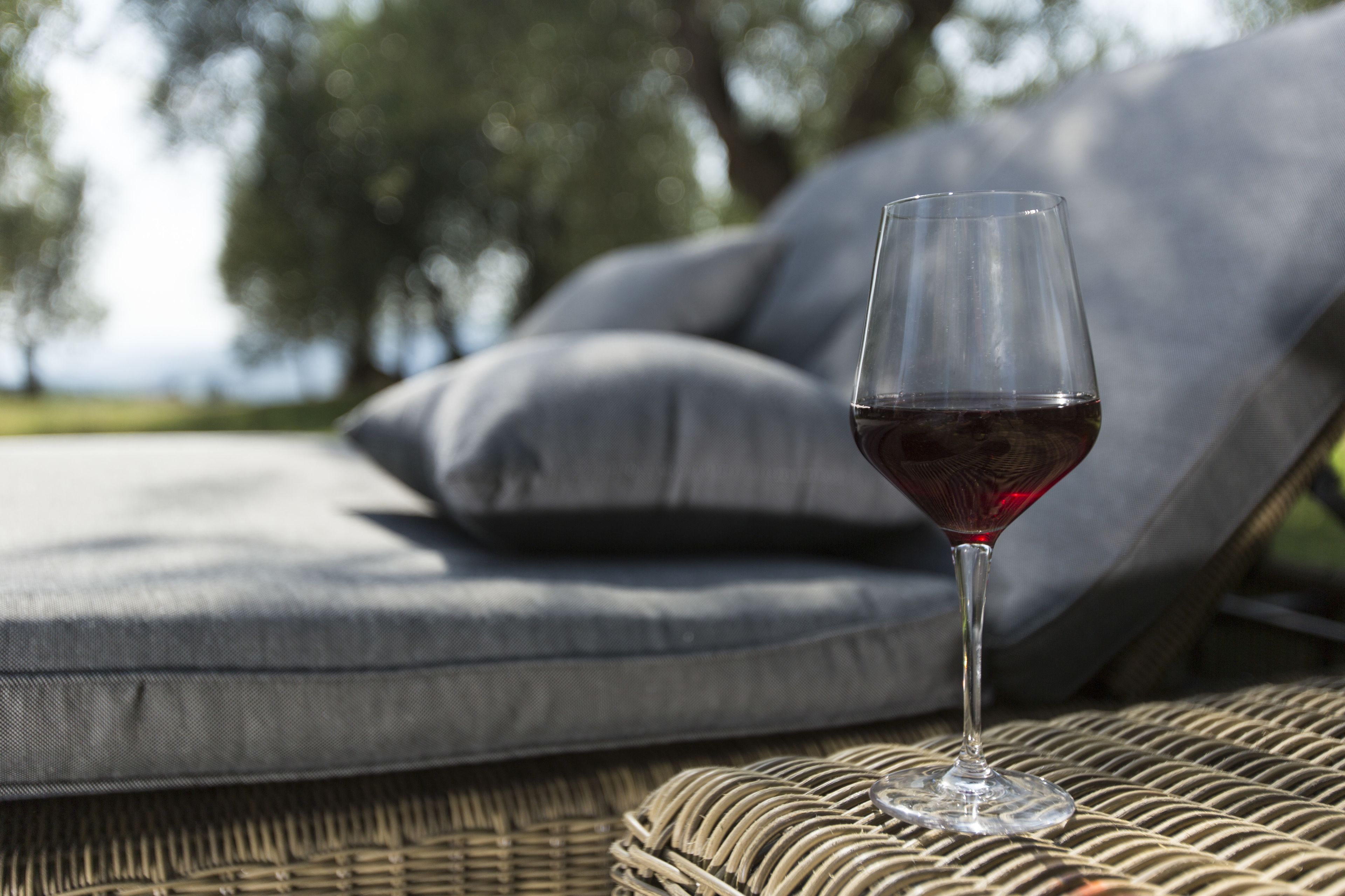 Un verre de vin rouge sur une table en osier, avec des coussins gris en arrière-plan.