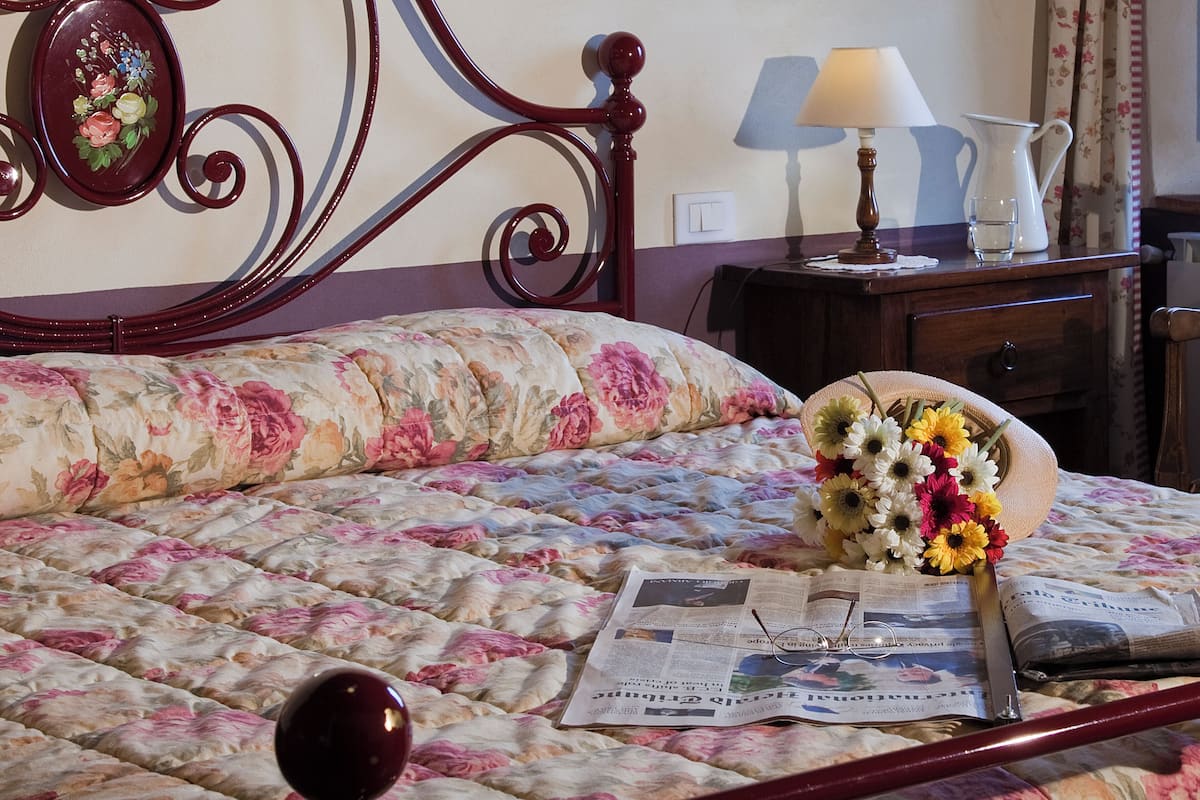 Een net opgemaakt bed met bloemenbeddels, een nachtkastje met een lamp, een bloemenvaas en een krant.
