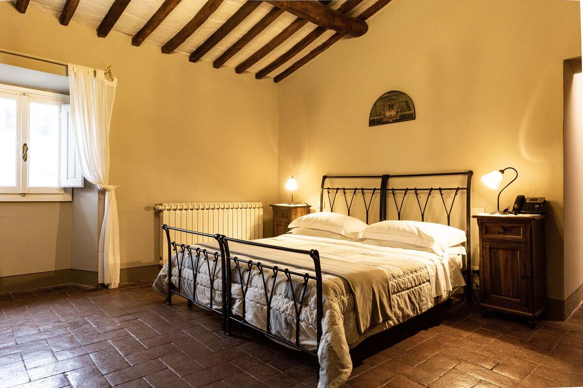 Een slaapkamer met een metalen bedframe, een houten kast, een nachtkastje en een raam met gordijnen.