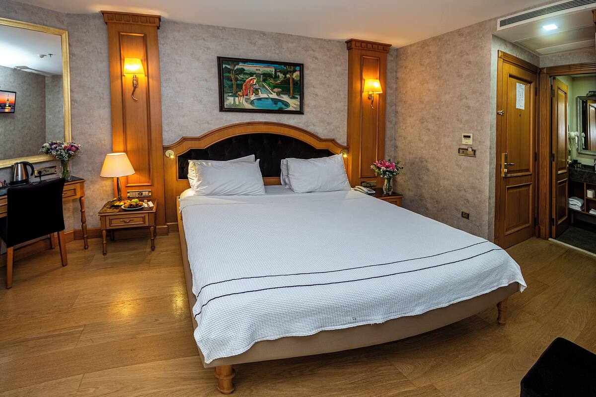 A hotel room with a large bed, a desk, a chair, a mirror, and a painting on the wall.