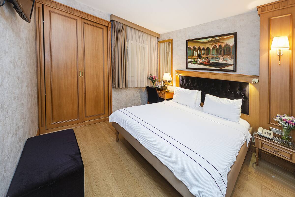 A hotel room with a large bed, wooden wardrobe, a nightstand with a lamp, and a painting on the wall.