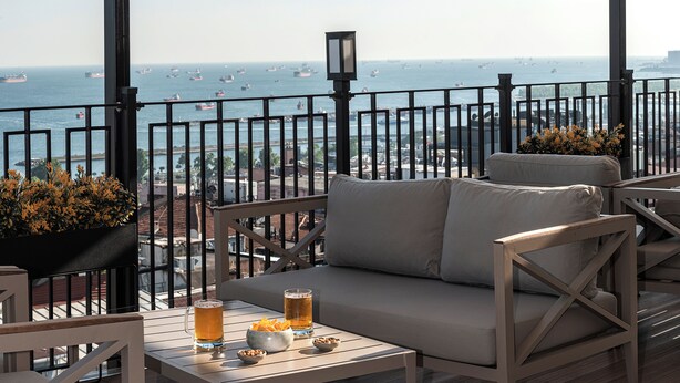 A balcony with a sofa, two tables with drinks and snacks, and a view of the sea with boats. - Aprilis Hotel - Special class (Istanbul)