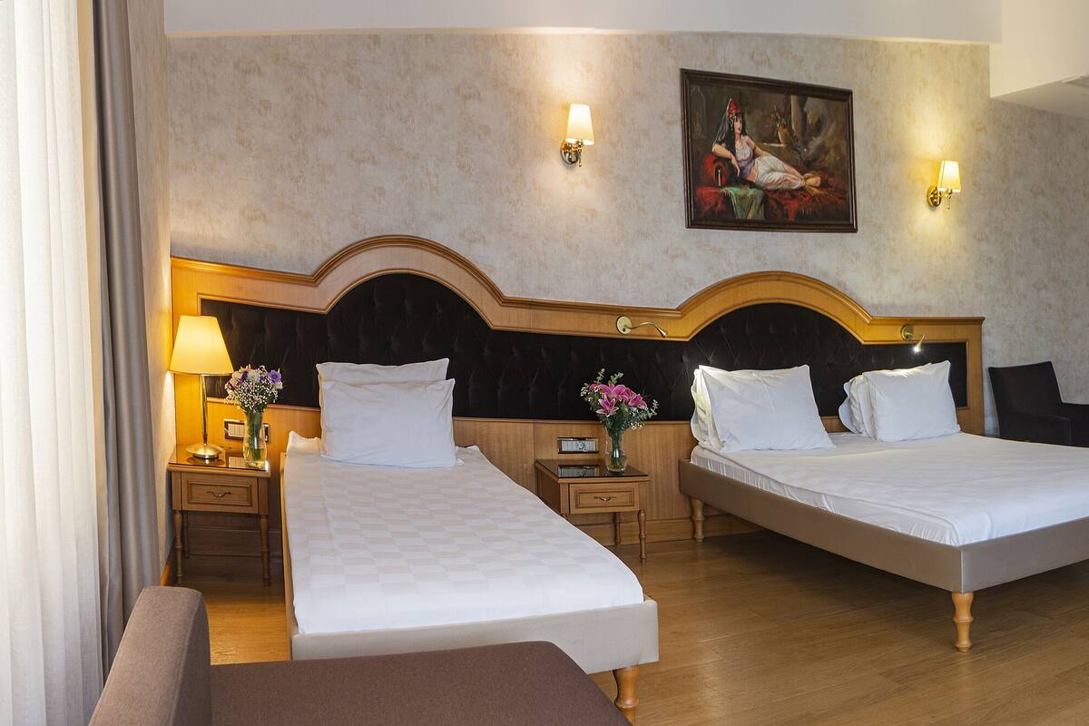 A hotel room with two beds, a wooden wardrobe, a desk with a chair, and a painting on the wall.