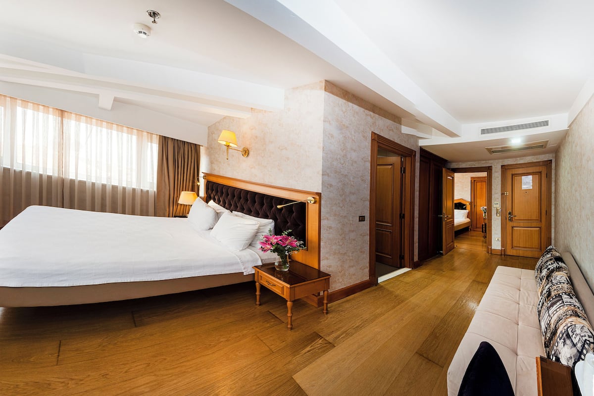 A spacious hotel room with a large bed, a bedside table with a lamp, a TV, and a view of the city through the window.