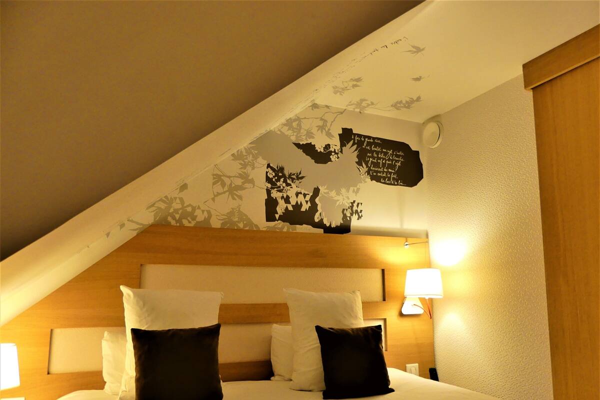 A hotel room with a sloped ceiling, a bed with white and dark pillows, and a wall with a floral mural.