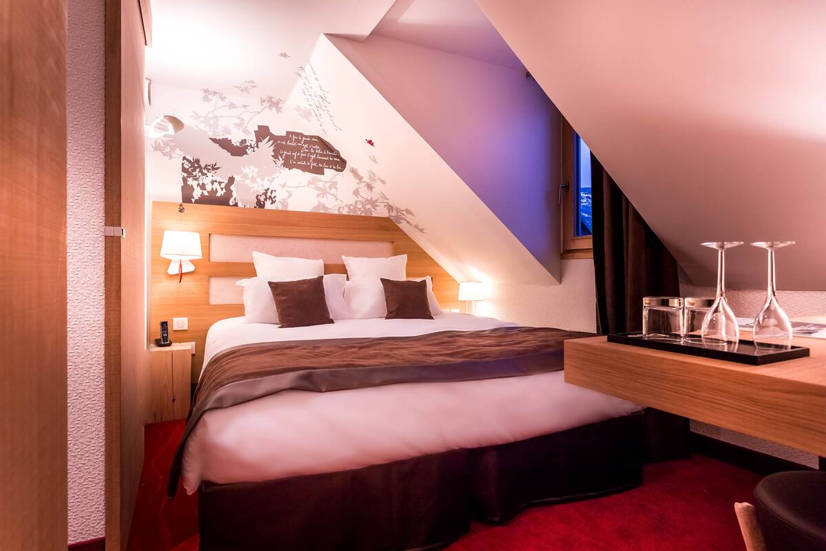 A modern hotel room with a neatly made bed, a wooden nightstand with a lamp, a desk with a tray of glasses, and a wall with decorative elements.